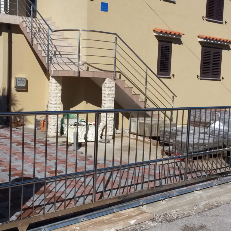 Metalna ograda i portuni od inoxa okružuju popločano dvorište s kamenim stupovima, vanjskim stubištem i bež zgradom sa zatvorenim tamnim kapcima i malim tendama od pločica iznad prozora.