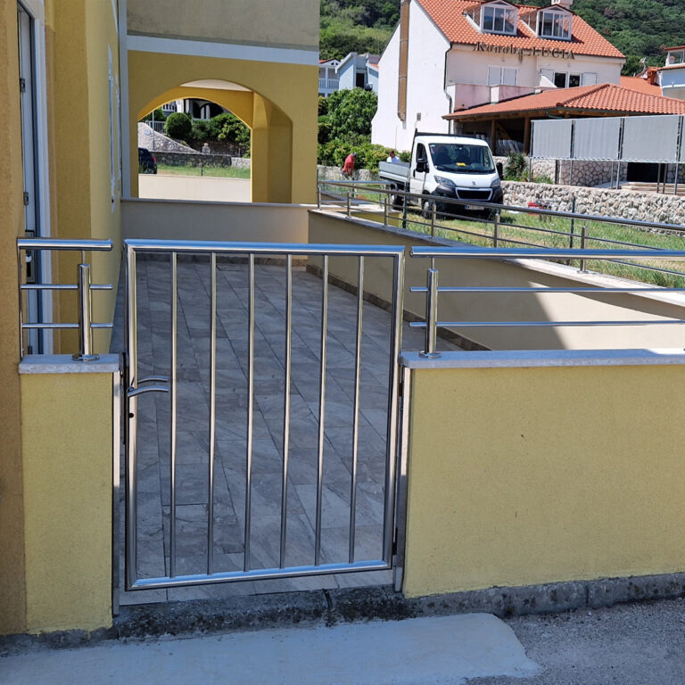 Zatvorena vrata od inoxa vode do popločane staze uz žutu zgradu. U pozadini je bijeli kombi parkiran pored kuće s crvenim krovom i drugih stambenih zgrada.