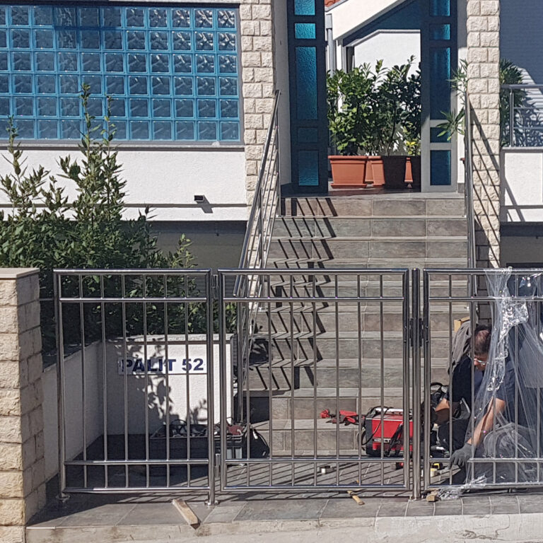 Metalna vrata od inoxa vode do stepenica ispred moderne kuće sa staklenim ciglama i biljkama u teglama. Dvoje ljudi rade s alatima i opremom blizu vrata za sunčanog dana.