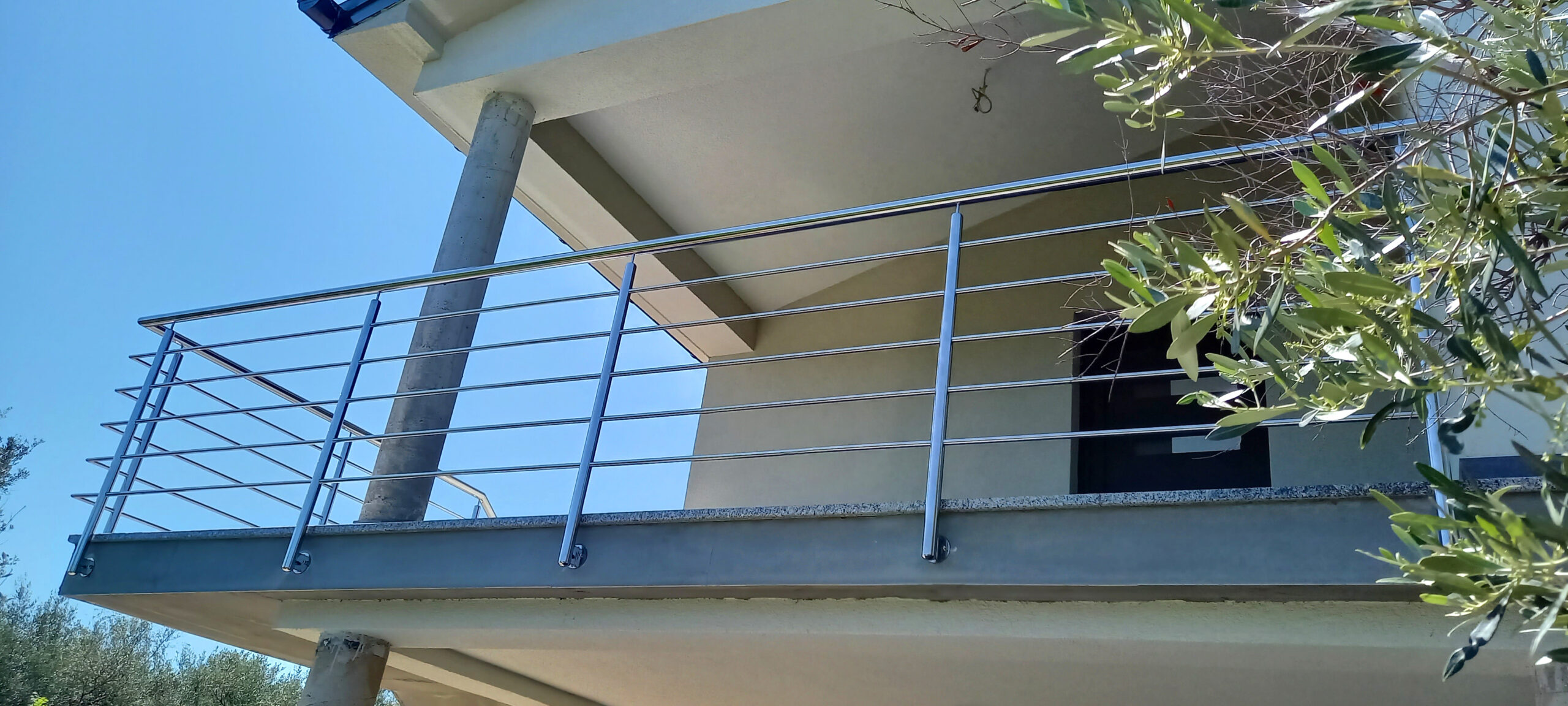 Moderni kućni balkon s ogradom od nehrđajućeg čelika, betonskim stupovima i krovnim prevjesom, okružen zelenim lišćem i uz vedro plavo nebo.